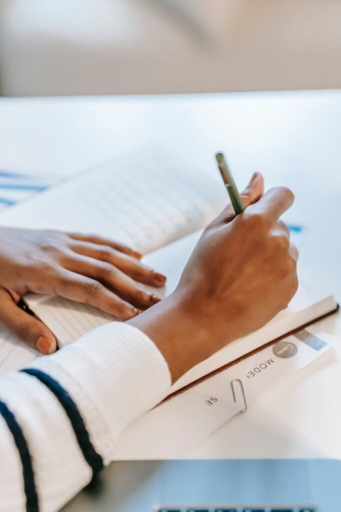 High angle of crop faceless ethnic female student writing information in notepad with pen on documents at white table in bright workspace